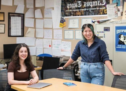 two students pose at a desk in front of The Campus Times poster and newspapers